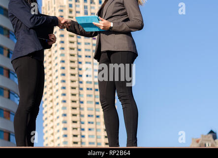 Two unidentifiable young women holding tablets shake hands with residential buildings in background suggesting a real estate or property transaction - Stock Photo