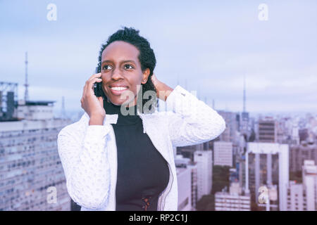 An African Brazilian woman on her mobile phone with the Sao Paulo skyline behind Stock Photo