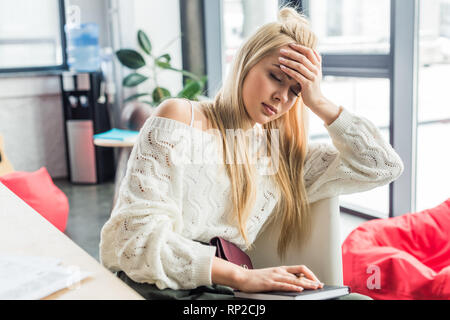 tired casual businesswoman sitting at desk with notebook and having headache in loft office Stock Photo