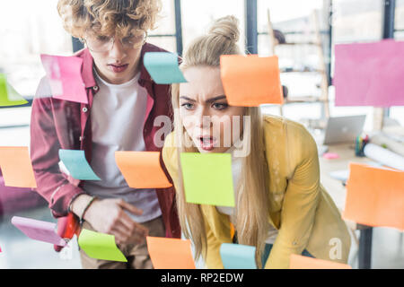 surprised casual businessman and businesswoman looking at colorful sticky notes on glass window in office Stock Photo