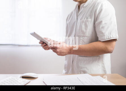Doctor in the uniform using computer tablet next to his office desk in hospital Stock Photo