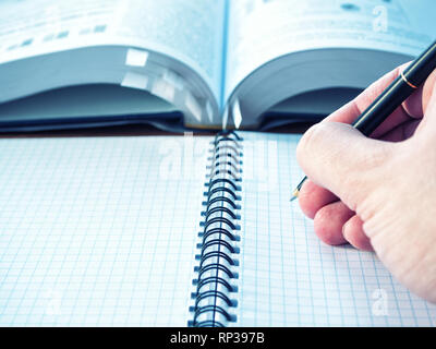 hand with pen above blank notepad pages with opened book blurred on background. close-up low angle image Stock Photo