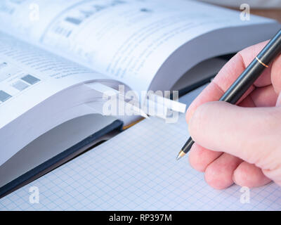 side view of a student hand with pen above empty sheets of squared notepad and blurred book with bookmarks on background. close-up low angle shot Stock Photo