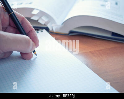 left handed student with pen preparing for exams, writing notes at textbook on wooden desk. opened encyclopedia dictionary with bookmarks on backgroun Stock Photo