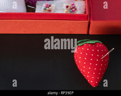 small needle pod in shape of strawberry with box full of needlework equipment on black background Stock Photo