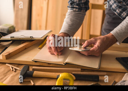 Carpenter is writing project notes while using smartphone in small business woodwork workshop Stock Photo