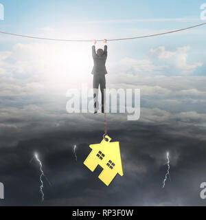 Businessman shackled by gold house hanging on the rope, on opposite weather conditions sky background, bright sun cloudscape and dark cloudy lightning Stock Photo