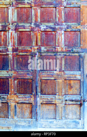 A door in a door! A large wooden door with square blocks, weathered with age and washed out, containing a smaller door with metal hinges and a latch. Stock Photo