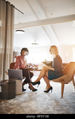 Two smiling young businesswomen discussing notes together and using a laptop while sitting in a modern office Stock Photo