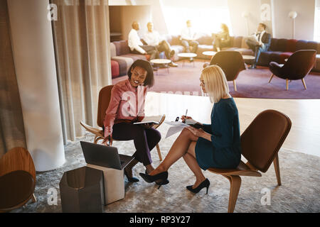 Two smiling businesswomen sitting in a modern office working on a laptop together and going over documents Stock Photo