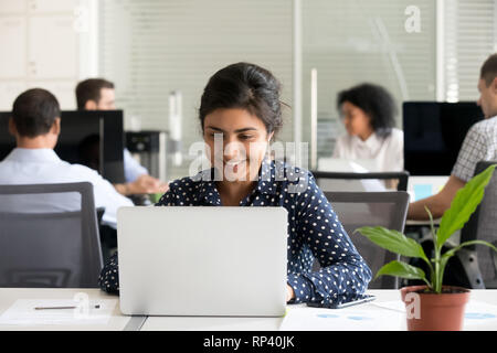 Indian worker sitting at desk reading business letter looking at computer screen feels happy and glad. Diverse employees sitting together in coworking Stock Photo