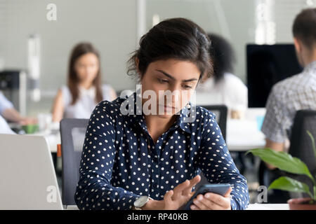 Indian concentrated worker sitting at desk in co-working office with diverse colleagues typing using cellular. Serious pensive employee receives unple Stock Photo
