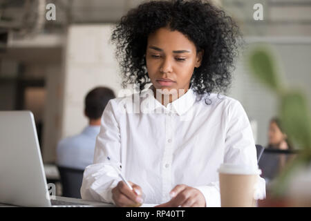 Serious millennial mixed race female sitting at desk holds pen making notes ideas in coworking space. African american pensive serious focused office Stock Photo