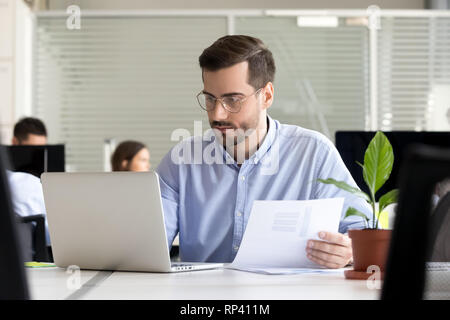 Serious thoughtful focused office worker manager sitting in coworking space office having busy day holds documentations sales statistics using compute Stock Photo