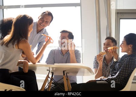 Diverse multiracial workers sitting around the office table holds slices of pizza eating junk food having pleasant conversation with coworkers. Positi Stock Photo