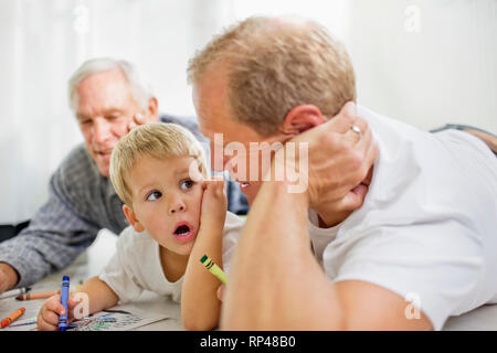 Young boy with his mid-adult father and mature grandfather lying on the floor coloring in. Stock Photo