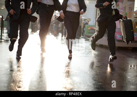 Four suited businesspeople running along a wet pavement in the city. Stock Photo
