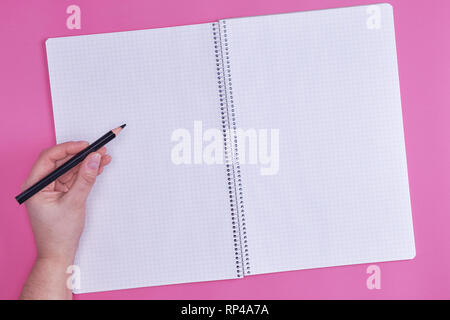 human hand holds black wooden pencil over empty open notebook, top view, pink background Stock Photo