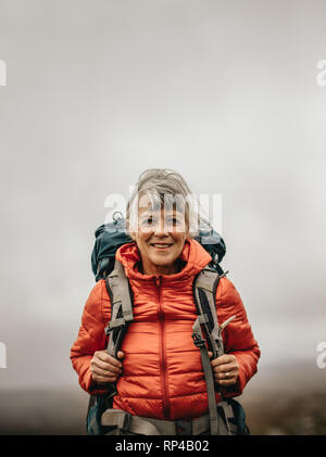 Smiling woman hiker wearing jacket and backpack on a hiking trip. Adventure seeking woman walking on a hill on a cloudy day. Stock Photo