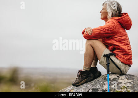 Side view of a female hiker sitting on a hill enjoying the view from the top. Senior woman sitting relaxed on a rock after her trek. Stock Photo