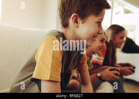 Group of kids having fun playing video game at home. Close up of a smiling boy sitting beside two girls playing video game holding joysticks. Stock Photo