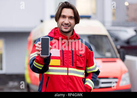 Smiling paramedic in red uniform holding smartphone with blank screen Stock Photo