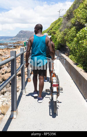 African man pushing a bicycle on a coastal footpath and walking in the hot Summer sun at Muizenberg, Cape Peninsula, South Africa Stock Photo