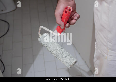 hand of working man in white, standing in empty room, holding paint roller . Painting apartment, renovating with white color paint Stock Photo