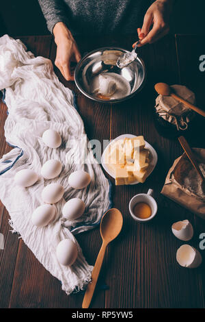 View from above young woman cooking dough for pie mixing sour cream, sugar and yolk in metal bowl next to eggs, diced butter and flour on brown wooden Stock Photo