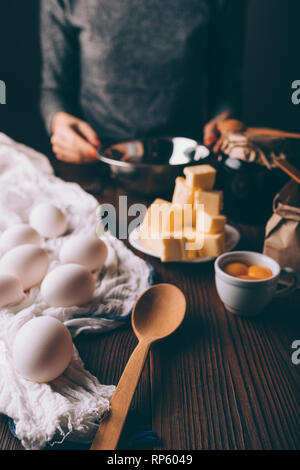 Process of making homemade cake on brown wooden table. Unrecognizable woman holding metal bowl near baking ingredients: eggs, yolks, diced butter and  Stock Photo