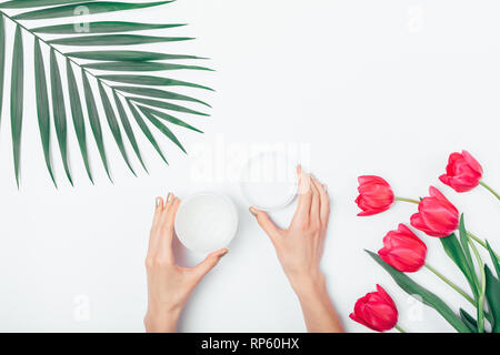 Woman's hands holding an open jar of cosmetic cream next to bouquet of pink tulips and palm leaf, top view. Stock Photo