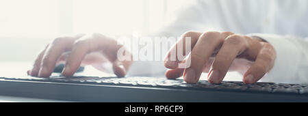 Wide view image of computer programmer typing using black keyboard with sun flare coming from the office window. Stock Photo