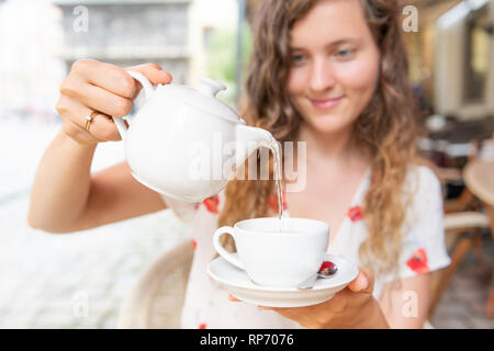 Young girl woman sitting in European outdoor cafe restaurant pouring green tea in white cup in summer in Lviv or Lvov, Ukraine city Stock Photo