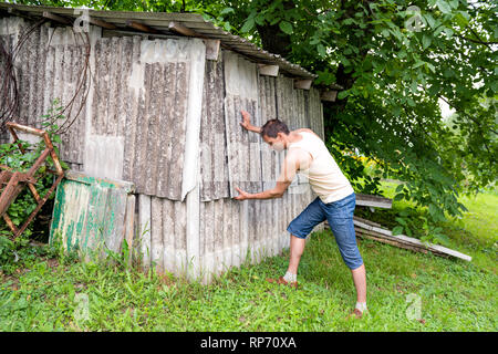Young man farmer in garden standing fixing building shed in green summer in Ukraine gray slate wall Stock Photo