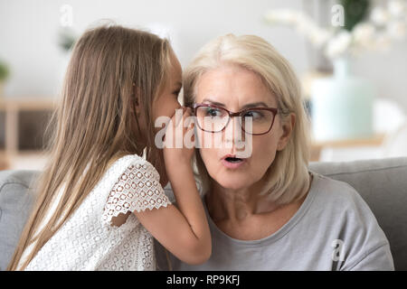 Little girl whispering in surprised grandmother ear, telling secret, grandma with wide open eyes listening to preschool granddaughter, family having f Stock Photo