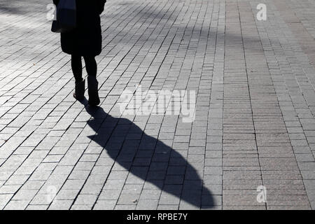 Slim girl walking down the street, black shadow and silhouette on sidewalk. Woman in warm clothes outdoor, concept of loneliness,  dramatic story Stock Photo