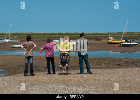 Four people standing on a shingle beach looking out over the tidal creeks  at Brancaster Staithe, North Norfolk, UK Stock Photo