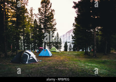 Camping tent on green grass beside lake with foggy over forest during sunrise. Tents on the edge of a forest in the mountains in the background. Stock Photo