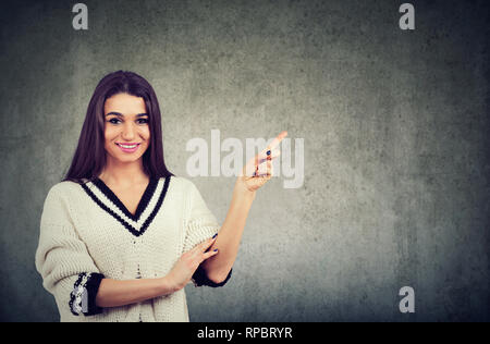 Portrait of a beautiful woman wearing white pullover expressing happiness pointing finger aside on copy space isolated over gray background Stock Photo