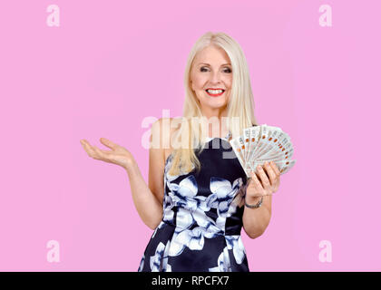 Happy, attractive blonde woman holding out a pile of ten pound notes, held in a fan shape and taken against a pink background Stock Photo