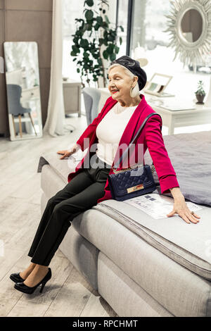 Beaming charming elderly lady happily sitting on new bed Stock Photo
