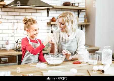 Beaming blonde mother in striped apron giving egg to curious daughter Stock Photo