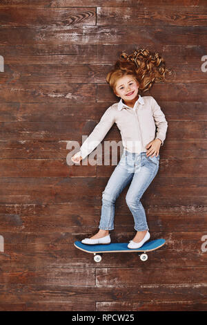 Happy child. Top view creative photo of little girl on vintage brown wooden floor. Girl on a skateboard, lying on the floor, looking at camera and smiling Stock Photo