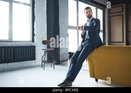 Waiting for a meeting. Serious handsome businessman weared in fashionable suit is looking at watch and waiting for important meeting. Fashion look. Business style. Stock Photo