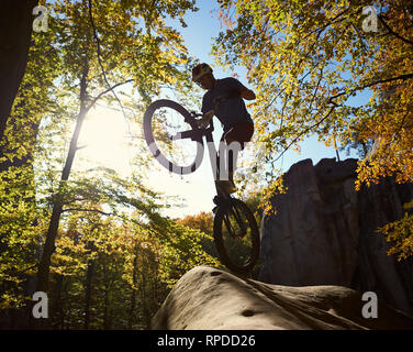 Sihlouette of professional sportsman cyclist standing on back wheel on trial bicycle, young man rider making acrobatic trick on the edge of boulder in the forest on sunny day. Concept of extreme sport