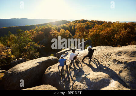 Back view of three young sportsmen with trial bicycles having a rest on the big boulder on the top of mountain. Guys enjoying beautiful view of setting sun and forest. Concept of extreme sport Stock Photo