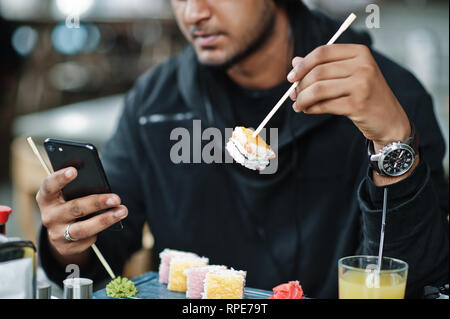 Close up photo of casual and stylish young asian man at cafe eating sushi and looking at mobile phone. Stock Photo