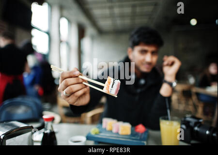 Close up photo of sticks holding casual and stylish young asian man with earphones at cafe eating sushi. Stock Photo
