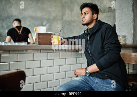 Casual and stylish young asian man with earphones at cafe drinking juice from glass. Stock Photo