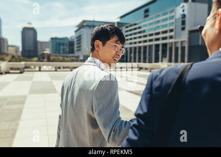 Rear view of happy asian businessman walking and talking with colleague outdoors in the city. Two businessmen talking and walking in the city. Stock Photo